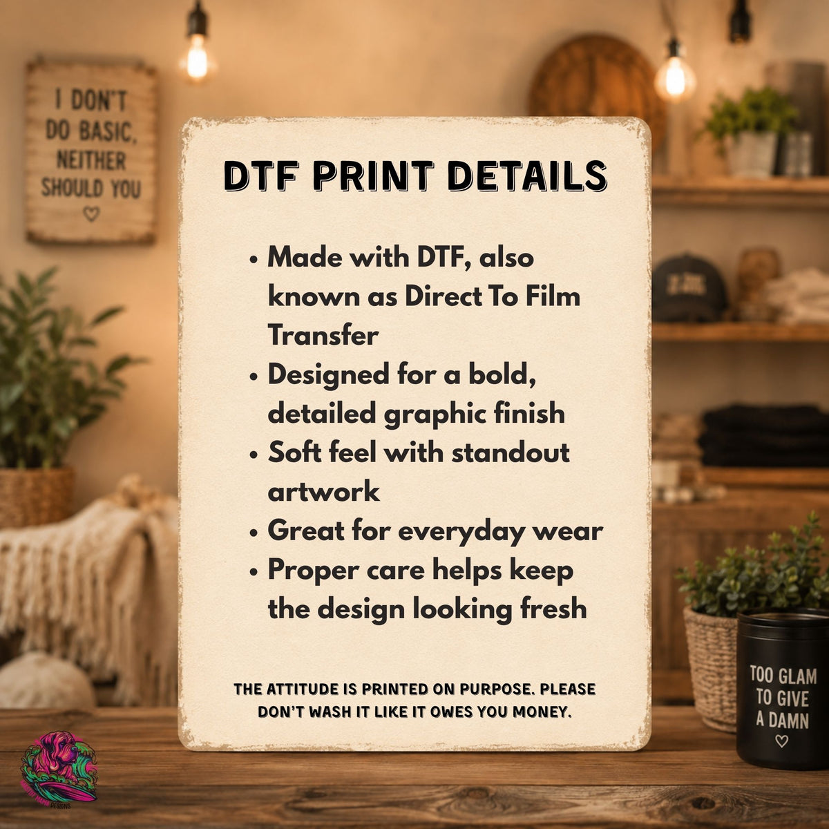 A rectangular printed sign with a distressed cream background and black text reading 'DTF PRINT DETAILS' with bullet points about Direct To Film Transfer, shown standing on a wooden table in a warm, styled room with plants and hanging bulbs.