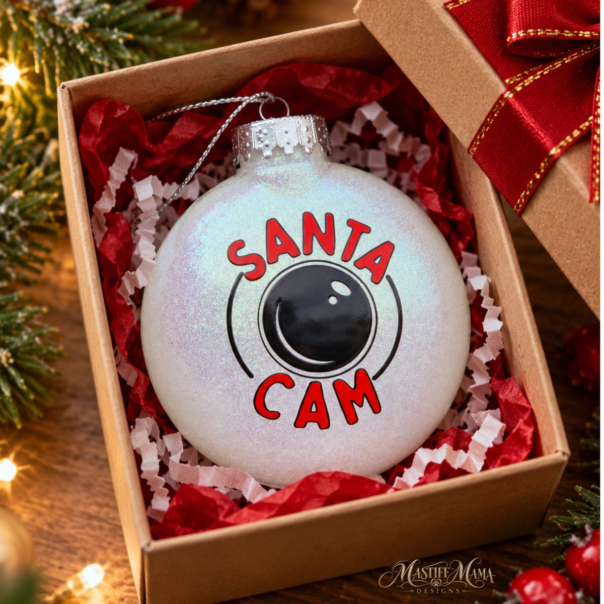 A round white glitter Christmas ornament with a Santa Cam badge design, displayed inside a brown gift box with red tissue paper and a red ribbon.