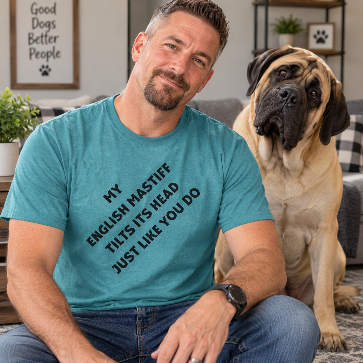 A man wearing a teal graphic t-shirt with black text that reads 'MY ENGLISH MASTIFF TILTS ITS HEAD JUST LIKE YOU DO' sits next to a large fawn and black Mastiff dog in a cozy living room setting.