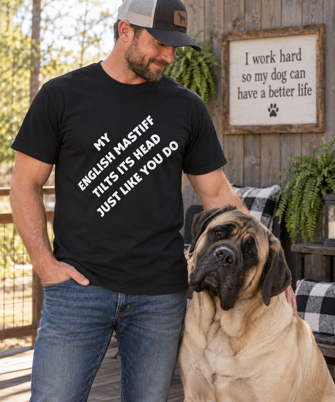 A man wearing a black graphic t-shirt with white text that reads 'MY ENGLISH MASTIFF THIS ITS HEAD JUST LIKE YOU DO' stands next to a large fawn and black dog on a wooden porch. A wooden sign on the background reads 'I work hard so my dog can have a better life'.