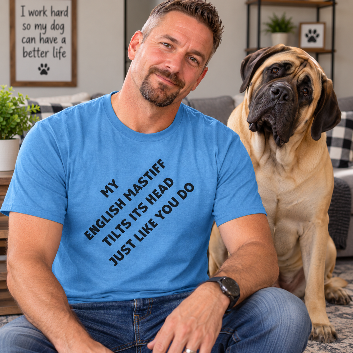 A man wearing a bright blue graphic t-shirt with the text 'MY ENGLISH MASTIFF TILTS ITS HEAD JUST LIKE YOU DO' sits next to a large tan Mastiff dog in a cozy living room setting.