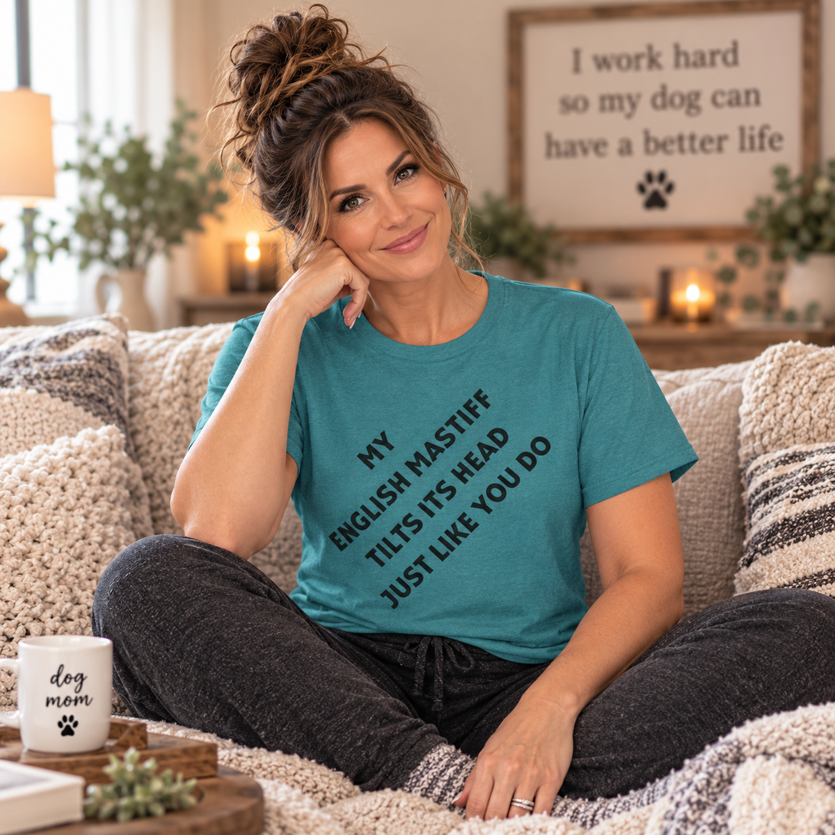 A woman sitting on a cozy couch wearing a teal graphic t-shirt that reads 'MY ENGLISH MASTIFF TILTS ITS HEAD JUST LIKE YOU DO'. She is wearing dark gray pants and sitting on a textured beige throw blanket in a warm, decorated living room.