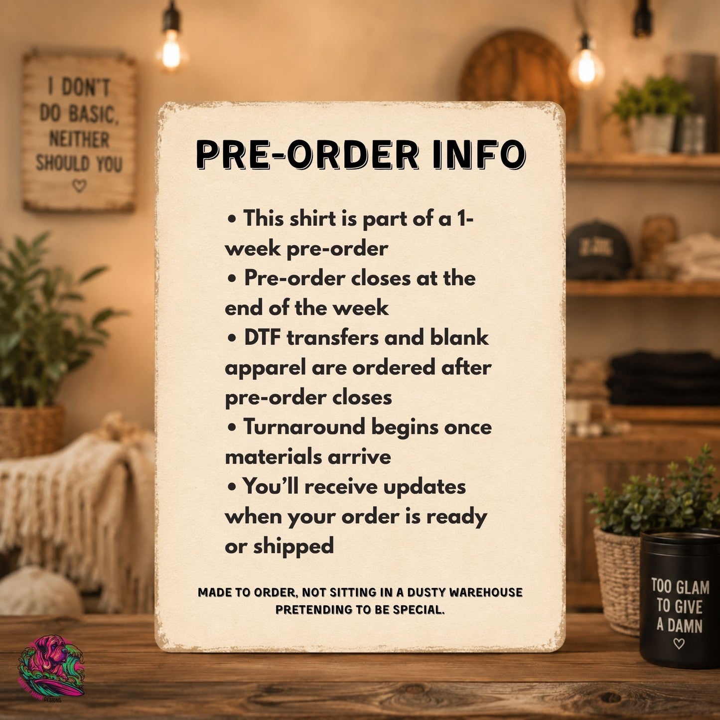 A rustic cream-colored sign with black text displaying 'PRE-ORDER INFO' and bullet points about a 1-week pre-order shirt, styled on a wooden table in a cozy, warm-toned room with plants and hanging lights.